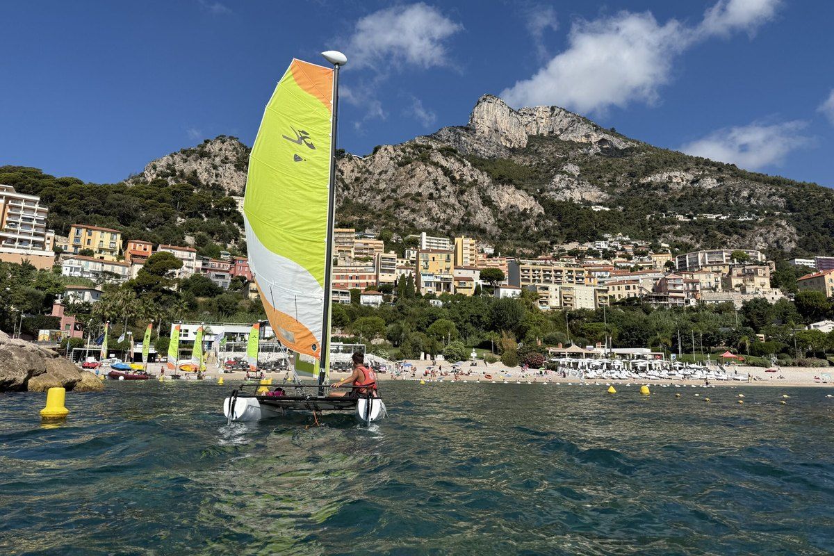 A small catamaran with a yellow and white sail off Cap-d'Ail, with the Tête de Chien rising behind