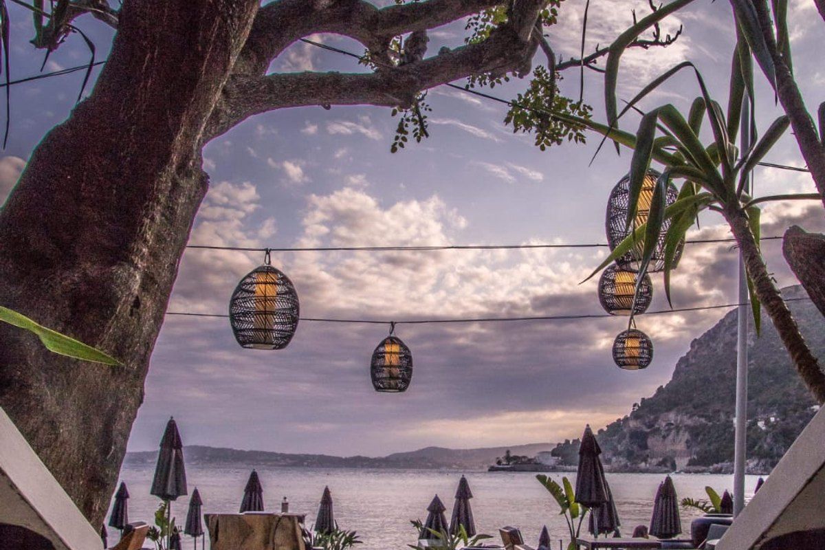 Lanterns strung between trees over the beach restaurant of Eden Plage Mala, with the cove and cliffs at dusk