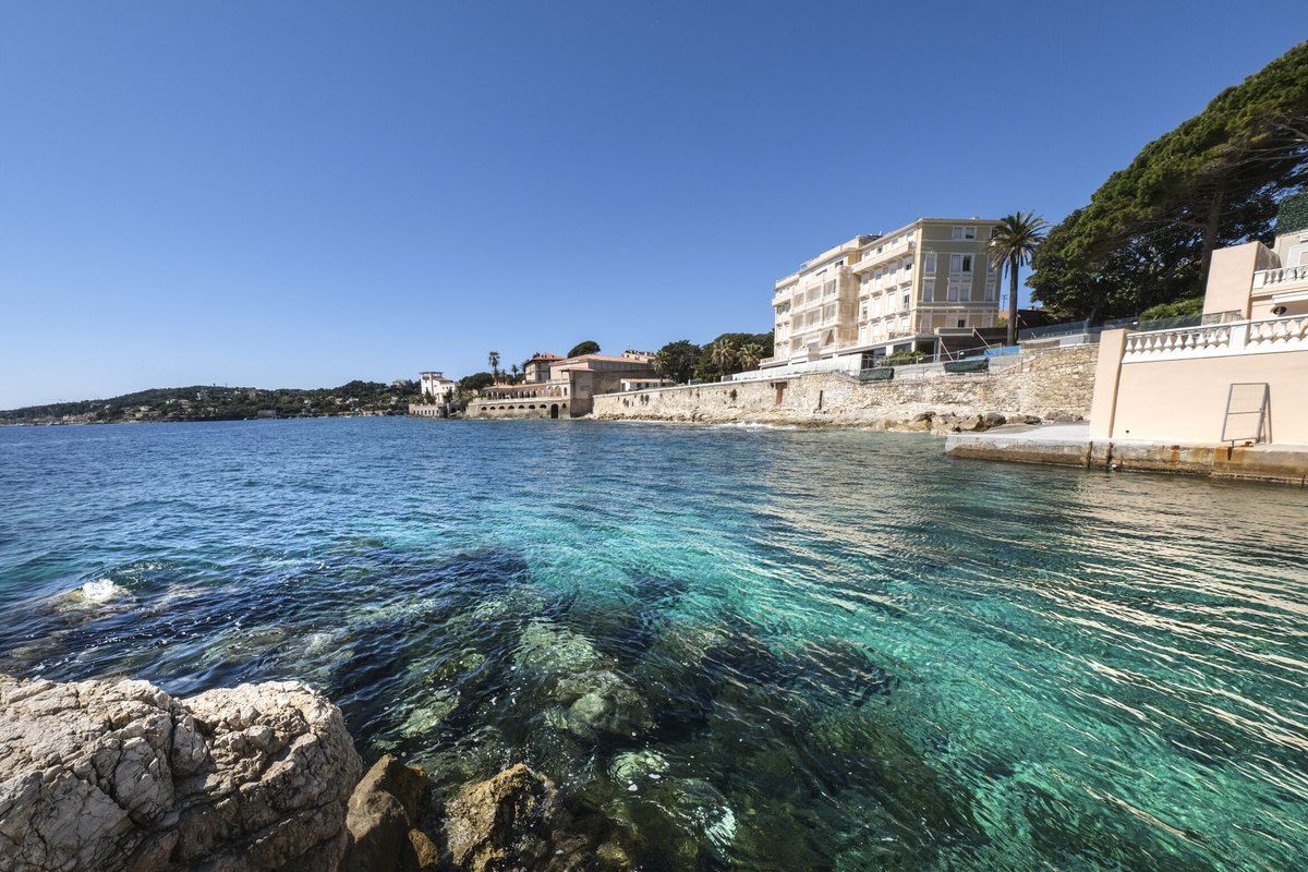 Clear green water over rocks beside the coastal promenade of Cap-d'Ail, with seafront buildings beyond
