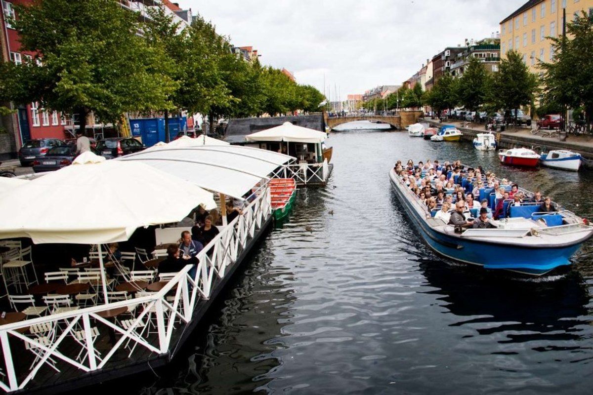 A low canal-tour boat full of passengers on Christianshavn Canal, with houseboats and waterside cafés alongside
