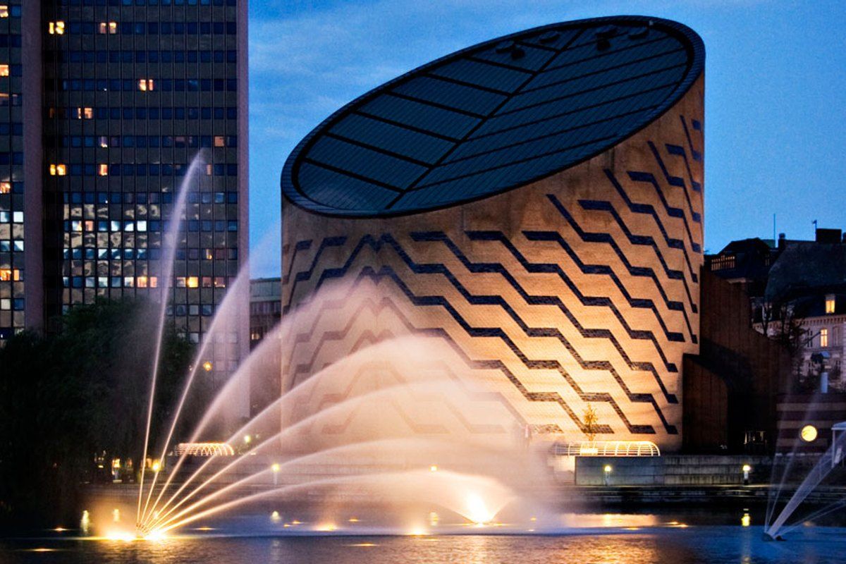 The cylindrical Tycho Brahe Planetarium lit up at dusk with fountains in Sankt Jørgens Sø in front