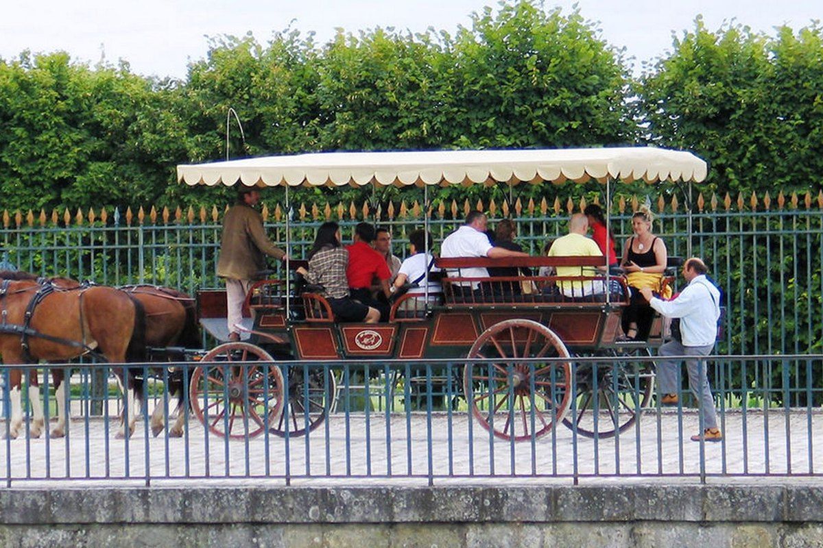 A two-horse open carriage with a striped canopy carrying passengers past the railings of the château park in Fontainebleau