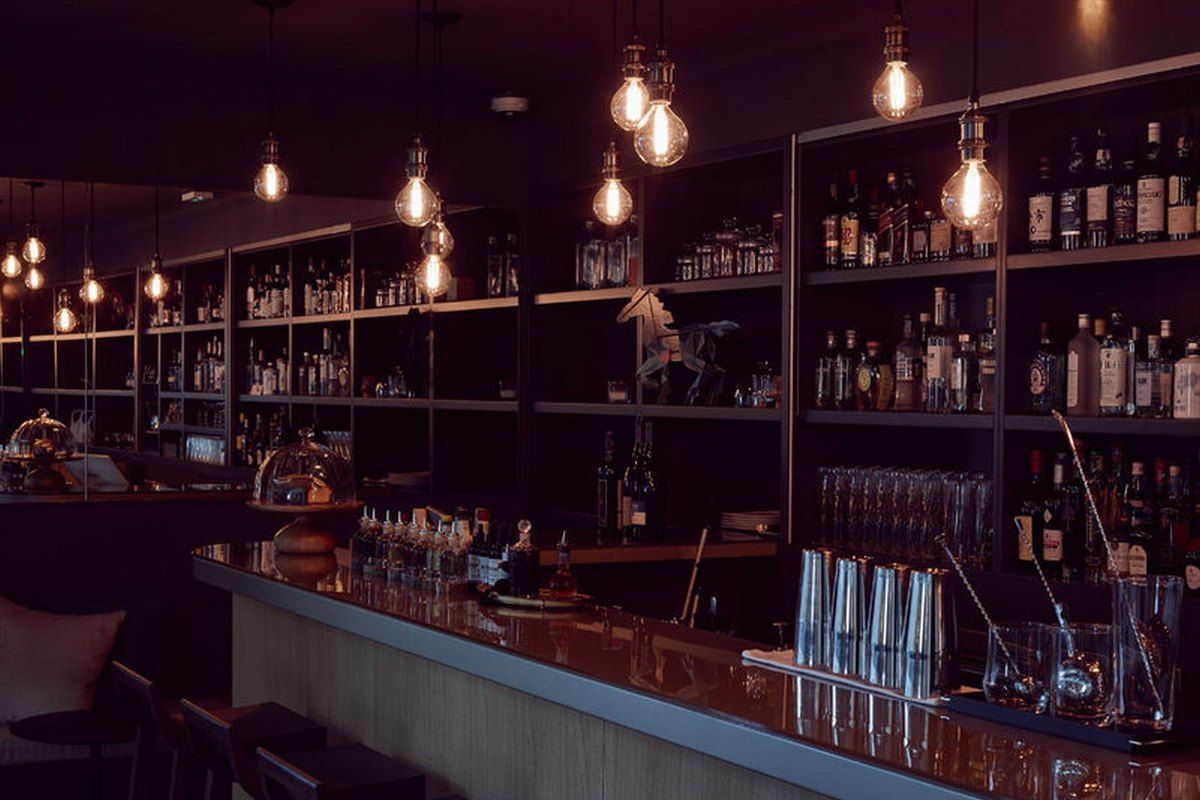 Backlit shelves of bottles and a long counter under hanging bulbs at Le Bar du Parc, Fontainebleau
