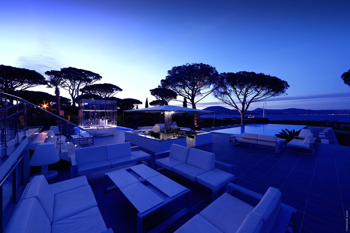 White lounge sofas on the sea-view terrace of the Kube Bar at dusk, umbrella pines beyond