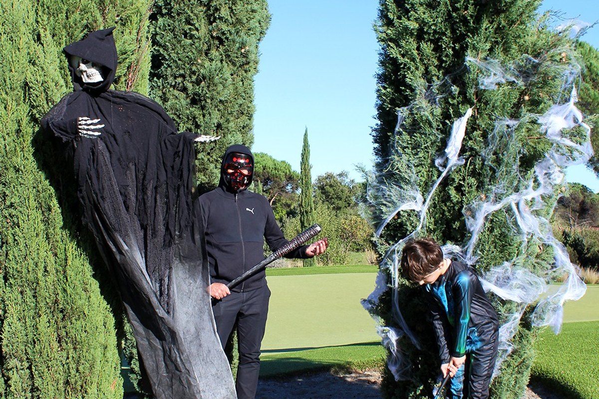 A grim reaper and a masked figure next to a young golfer on a putting green decorated with cobwebs for Halloween at Golf Up