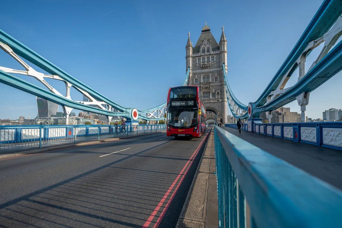 A red double-decker bus crossing Tower Bridge beneath the North Tower and blue suspension chains