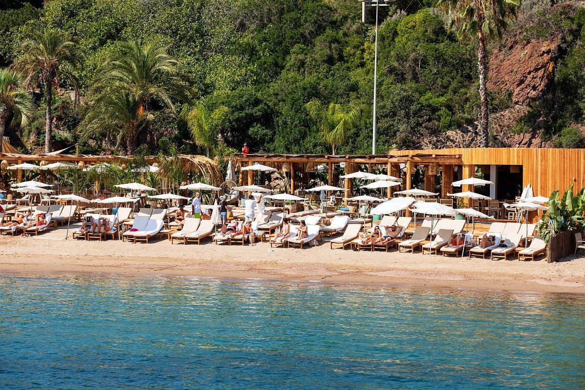 Loungers and parasols of Baia Plage on the sand at Port de la Rague, below green hills and red rocks