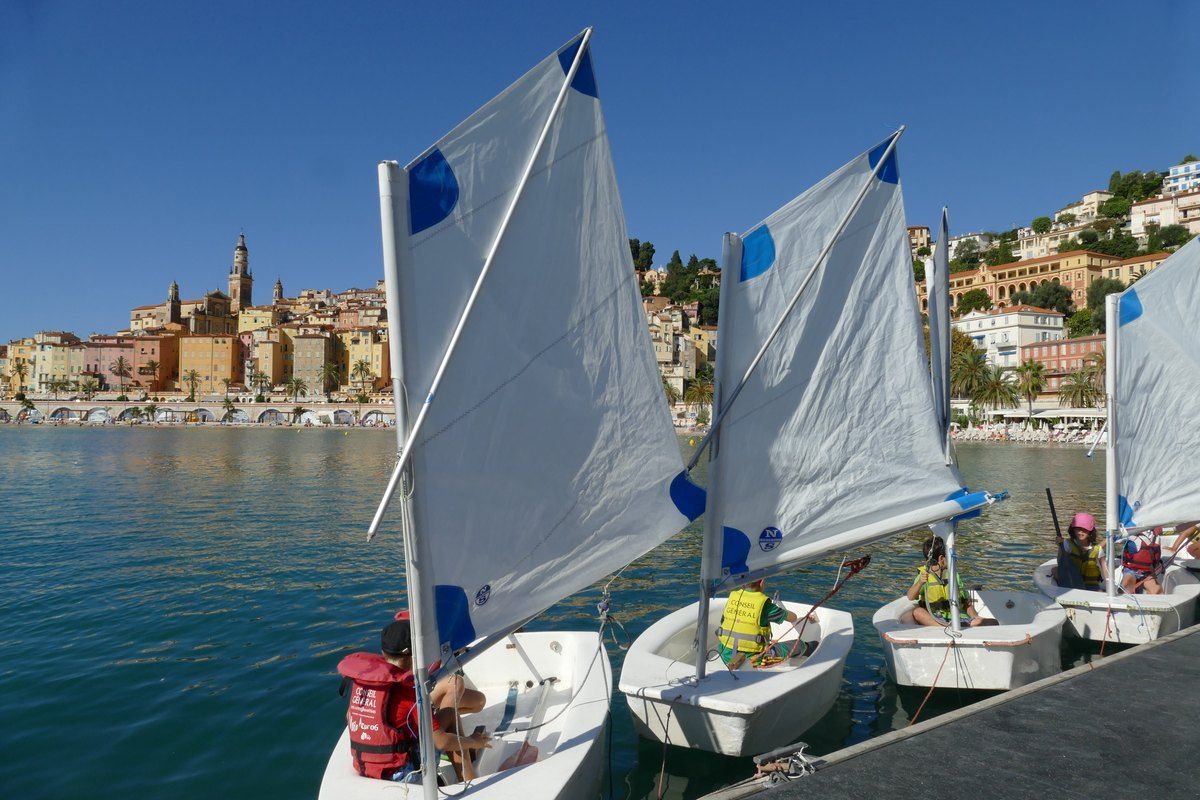 Children sailing Optimist dinghies off Menton, with the old town behind