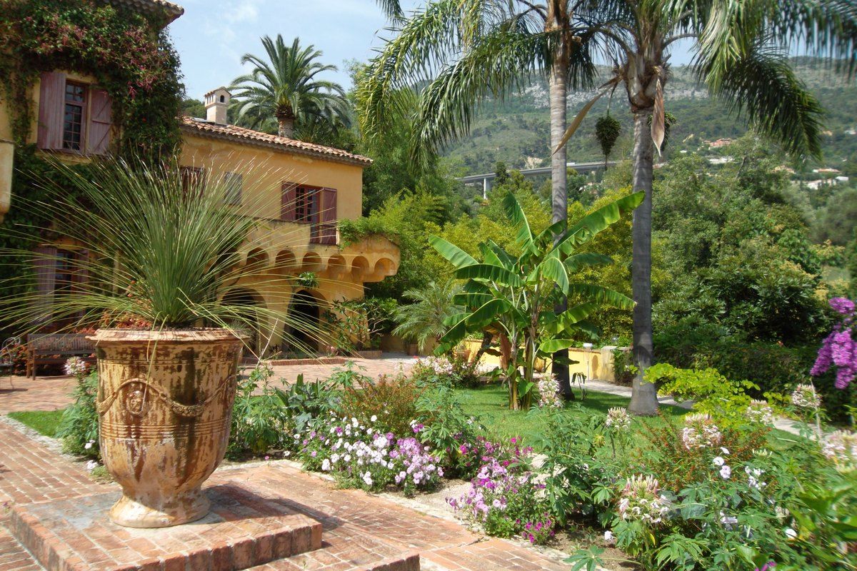 The ochre villa and flowering beds of the Val Rahmeh botanical garden in Menton, with palms and a terracotta urn