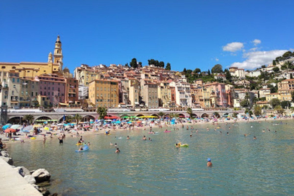 Plage des Sablettes in Menton with bathers in shallow water and the old town's pastel houses and basilica behind