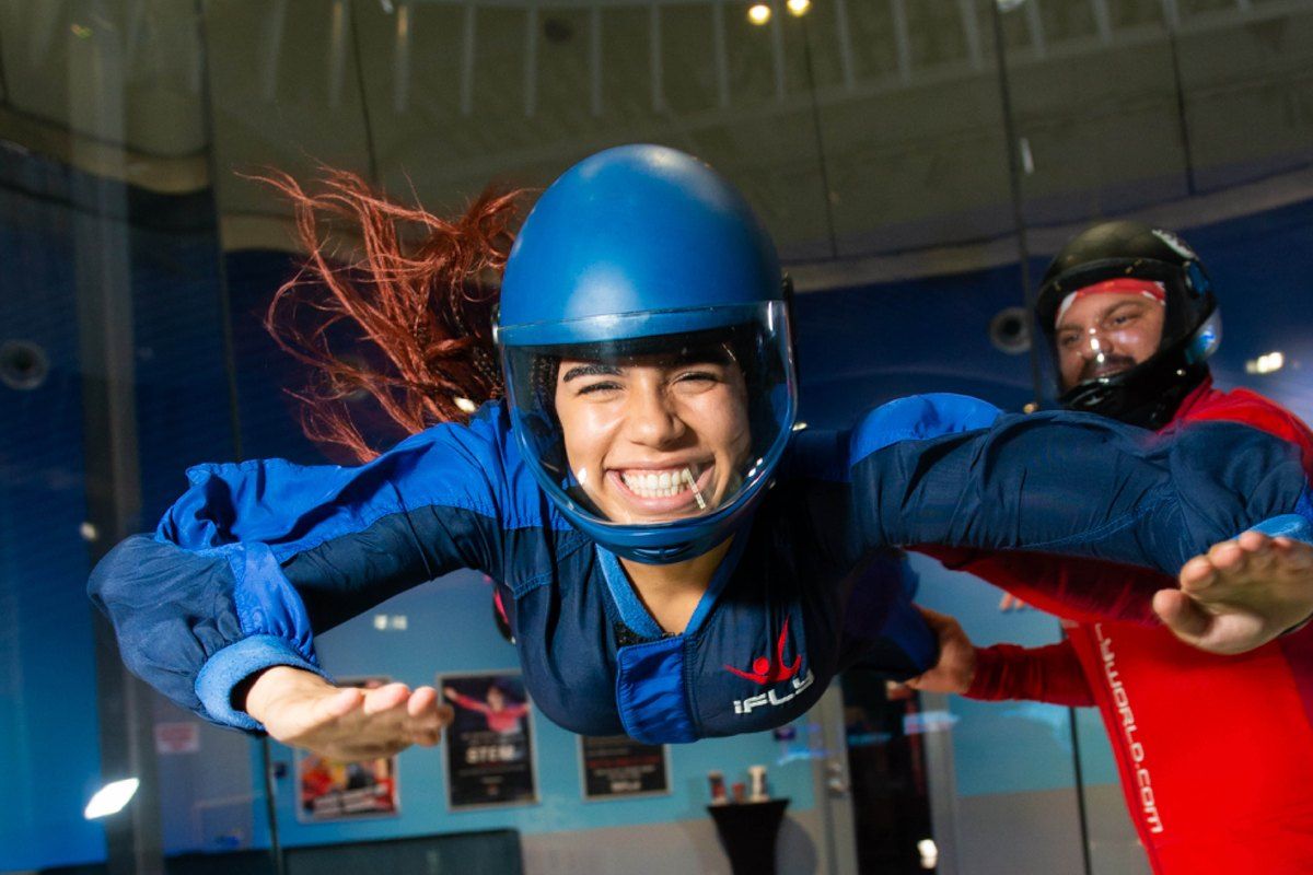 A smiling girl in a helmet and flight suit floating in the vertical wind tunnel at iFLY Miami with an instructor