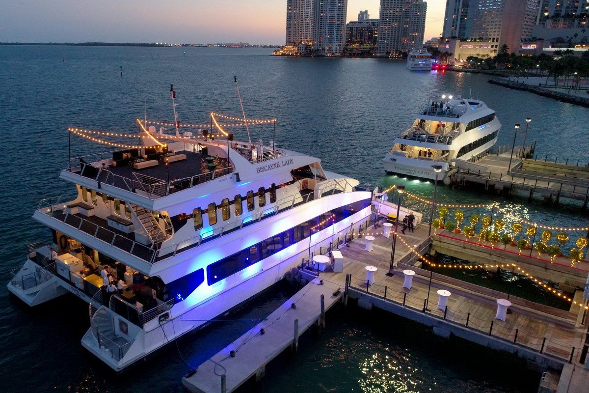 An Island Queen sightseeing yacht lit up at the Bayside dock at dusk, with the Downtown Miami skyline behind