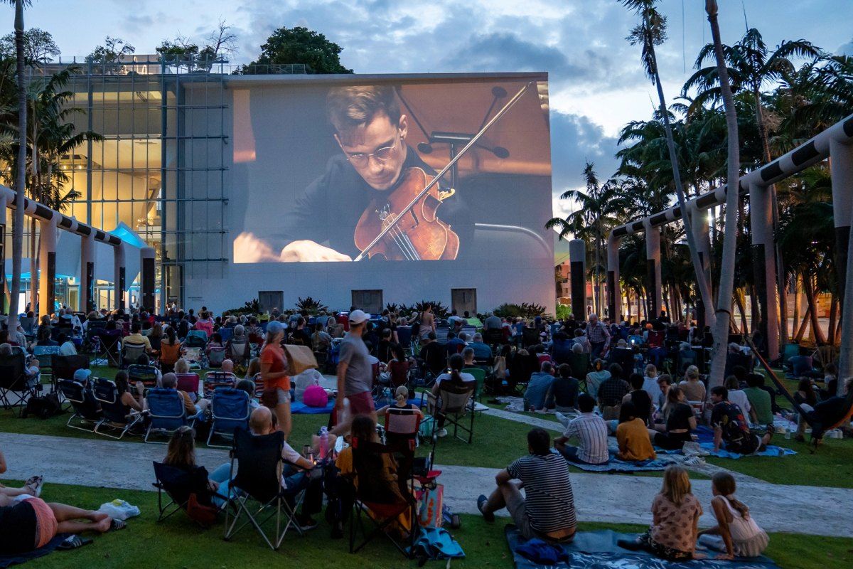 An audience on the lawn of SoundScape Park at dusk watching a violinist projected on the wall of the New World Center