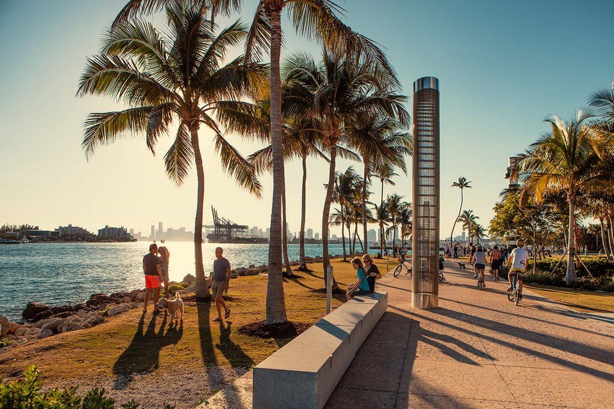 Walkers and cyclists on the palm-lined promenade of South Pointe Park in late-afternoon light, with the channel and PortMiami behind