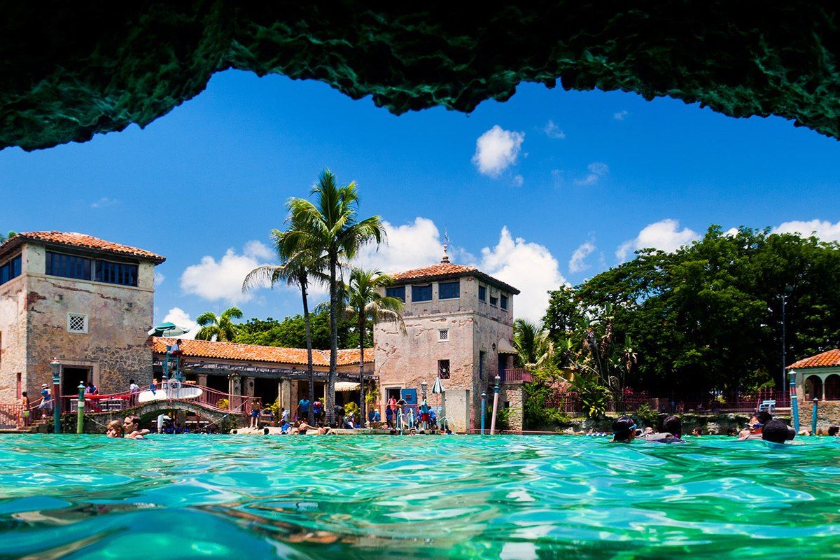 View from inside a coral-rock grotto across the turquoise water of the Venetian Pool to its loggias and towers