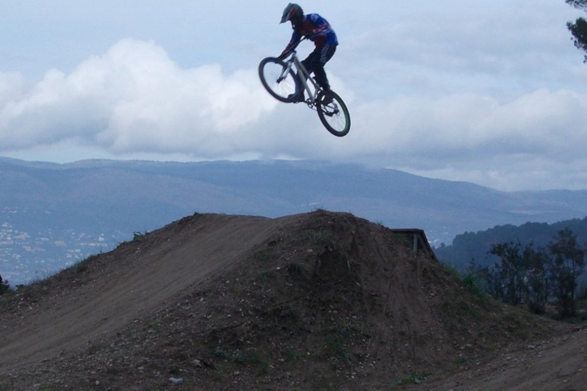 A mountain biker in mid-air over a jump at the Mougins bike park, with hills behind