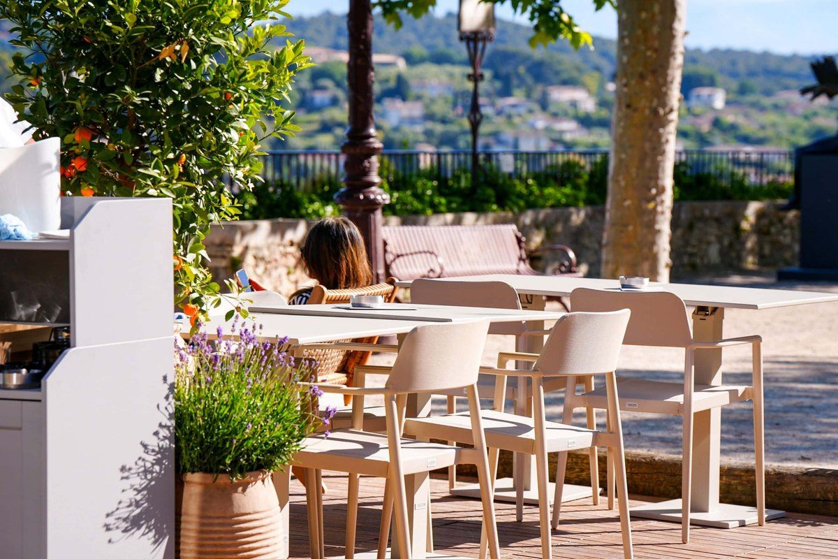 Sunlit terrace tables of the Café de la Place on Place des Patriotes, with the hills beyond the railing