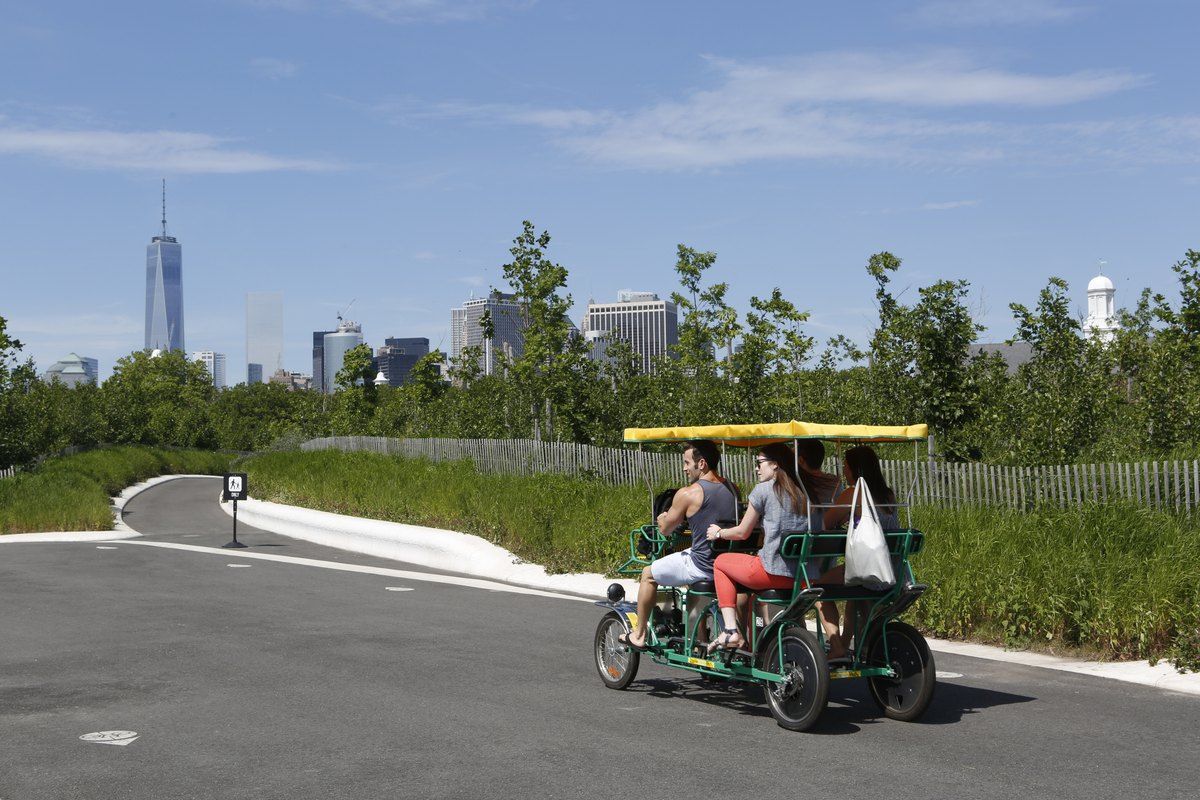 Visitors pedalling a surrey bike on Governors Island, with One World Trade Center across the water