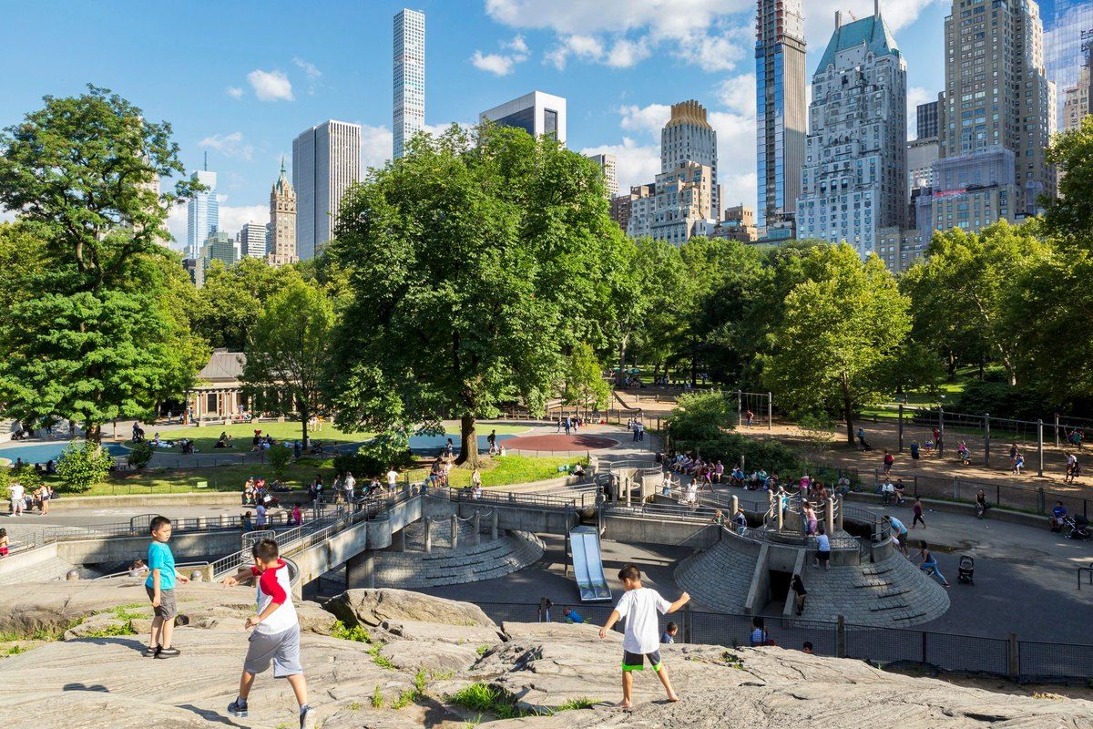 Children playing among the rocks and climbers of Heckscher Playground, with the Central Park South towers behind