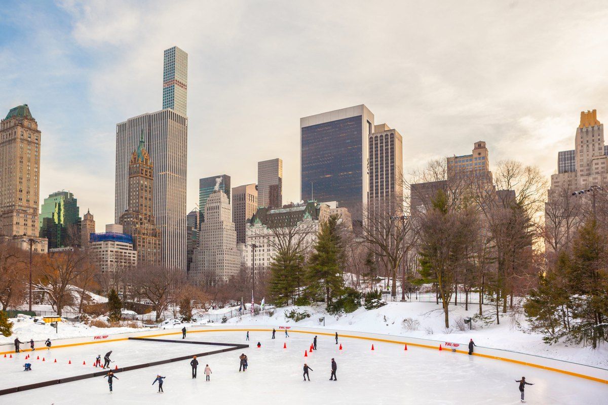 Skaters on Wollman Rink in a snowy Central Park, with the Midtown skyline behind
