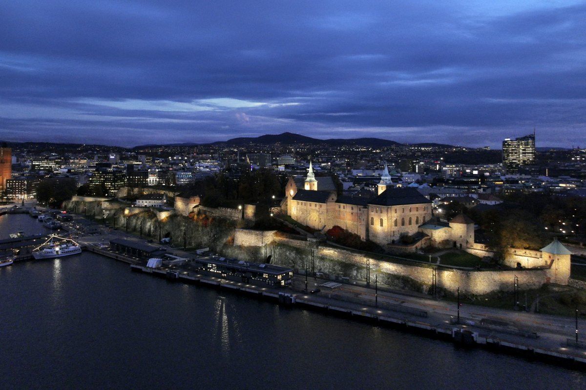 Akershus Fortress and castle lit up above the harbour at dusk, with Oslo behind