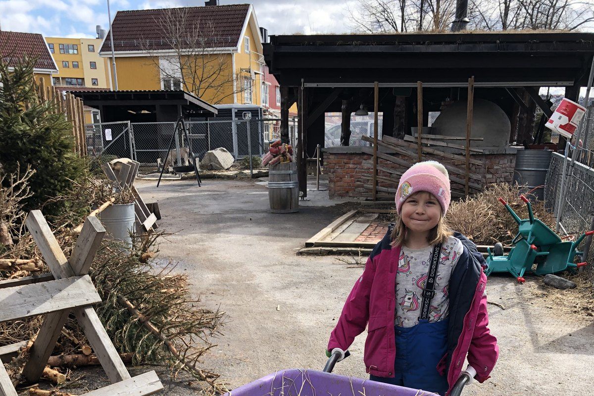 A smiling girl pushing a wheelbarrow across the yard at Kampen children's farm in Oslo