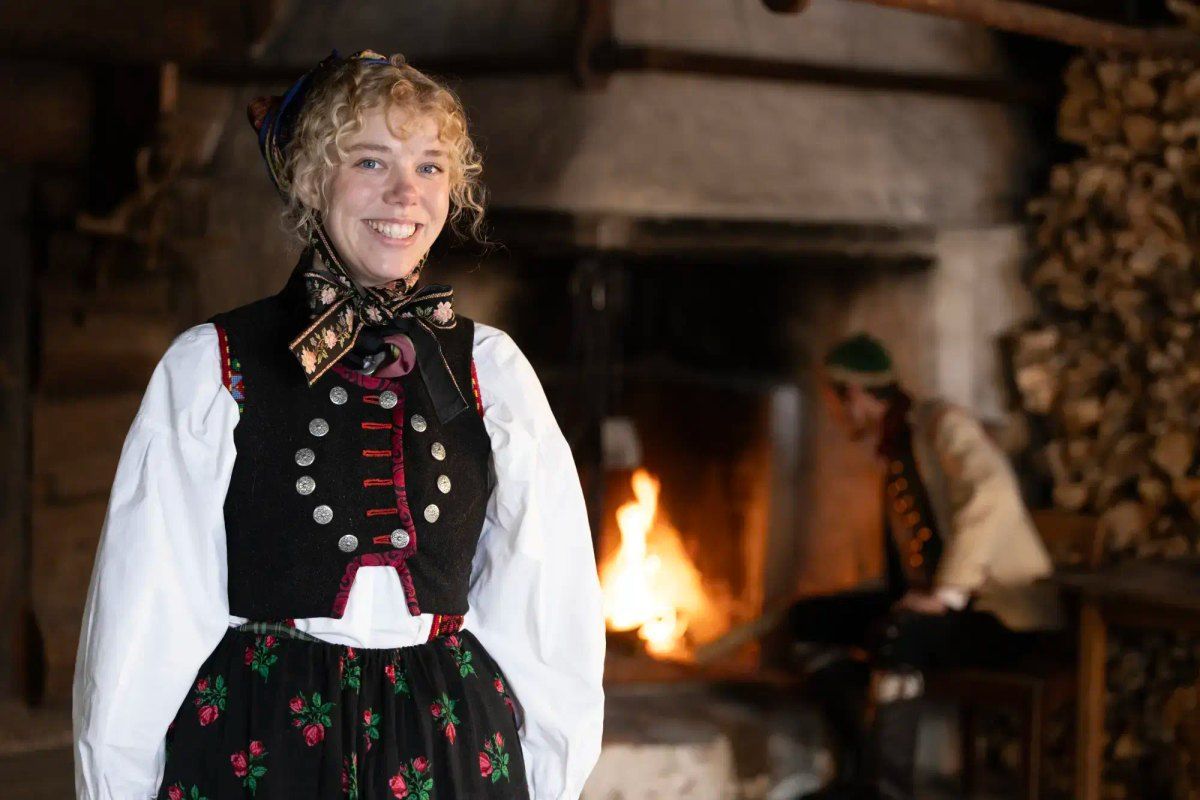 A host in Norwegian folk costume smiling beside an open fire in a log house at Norsk Folkemuseum, Bygdøy