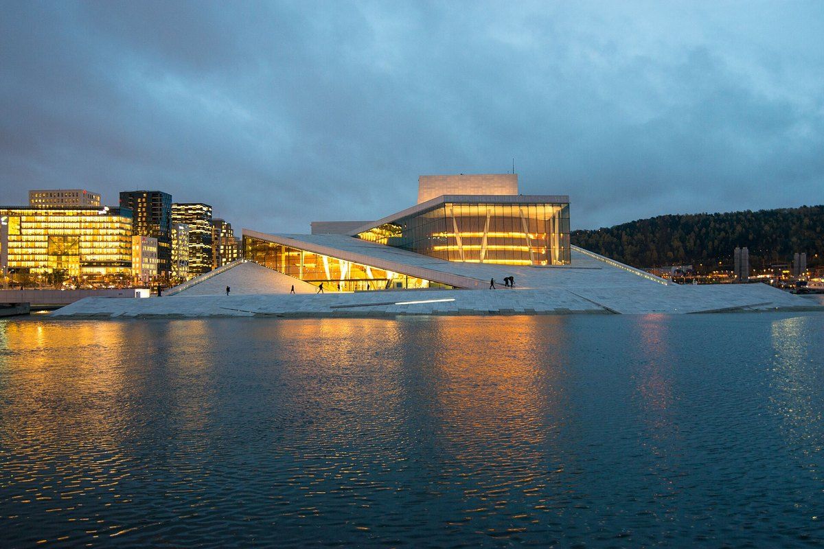 The Oslo Opera House glowing at dusk across the water, its white marble roof sloping down to the fjord