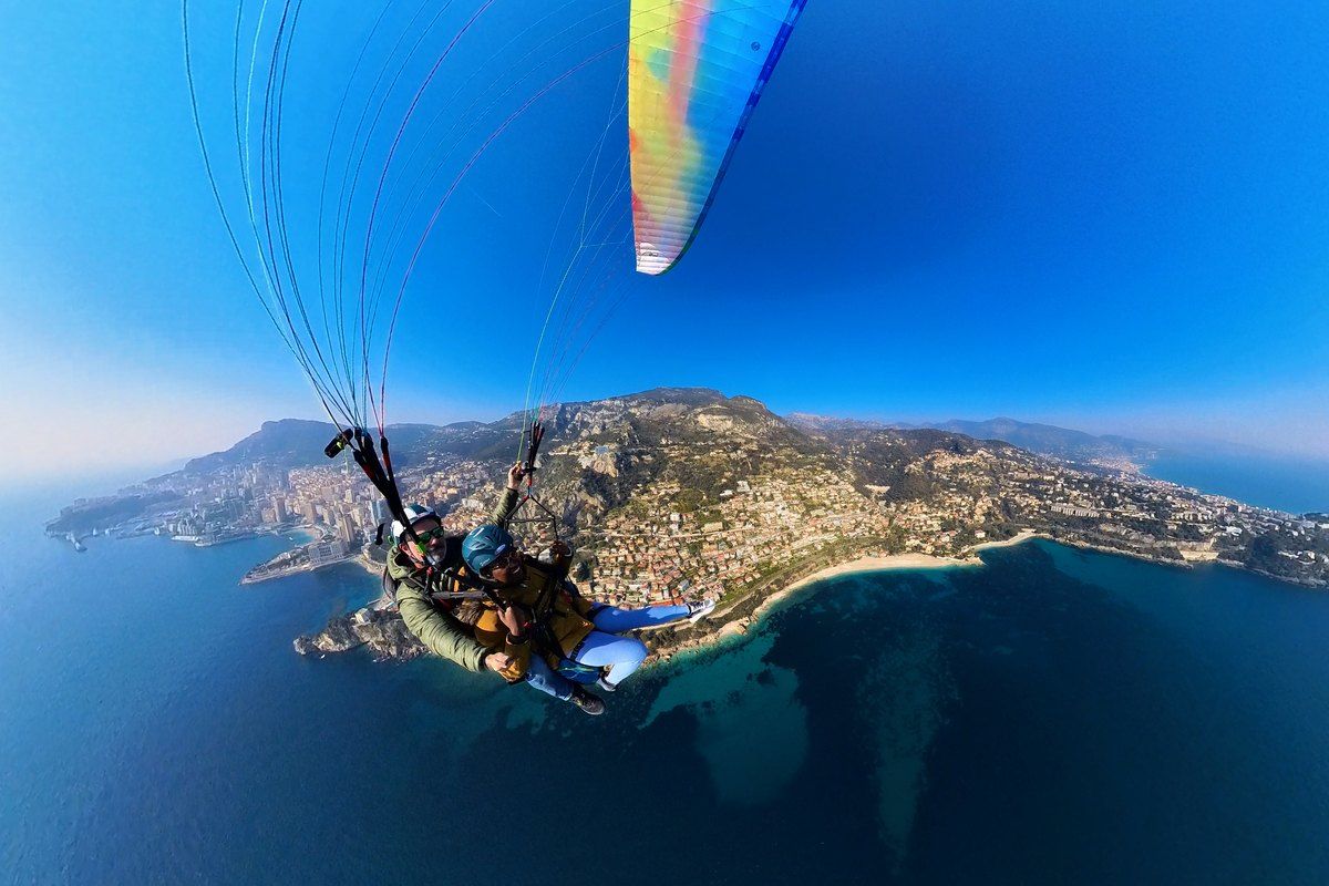 A tandem paraglider flying high above Cap Martin and the turquoise sea