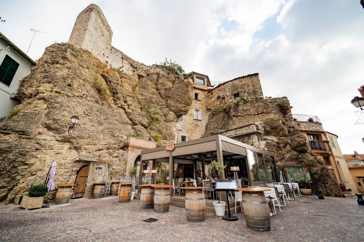 A café terrace with wine barrels on a cobbled square below the great rock and castle of Roquebrune village