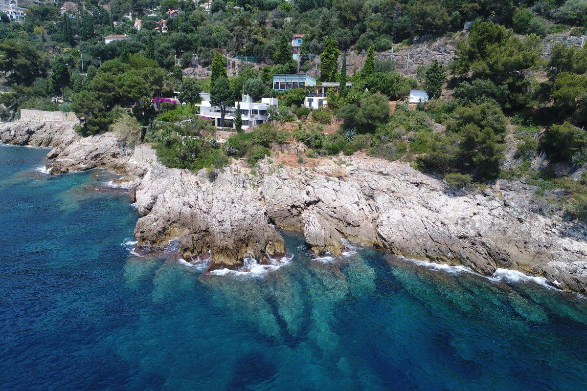 Drone view of the rocky shore and clear water below villas and pines along the Sentier Le Corbusier