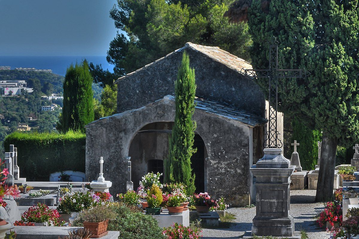 The small stone chapel in Saint-Paul de Vence cemetery among cypresses and flowers, with the sea beyond