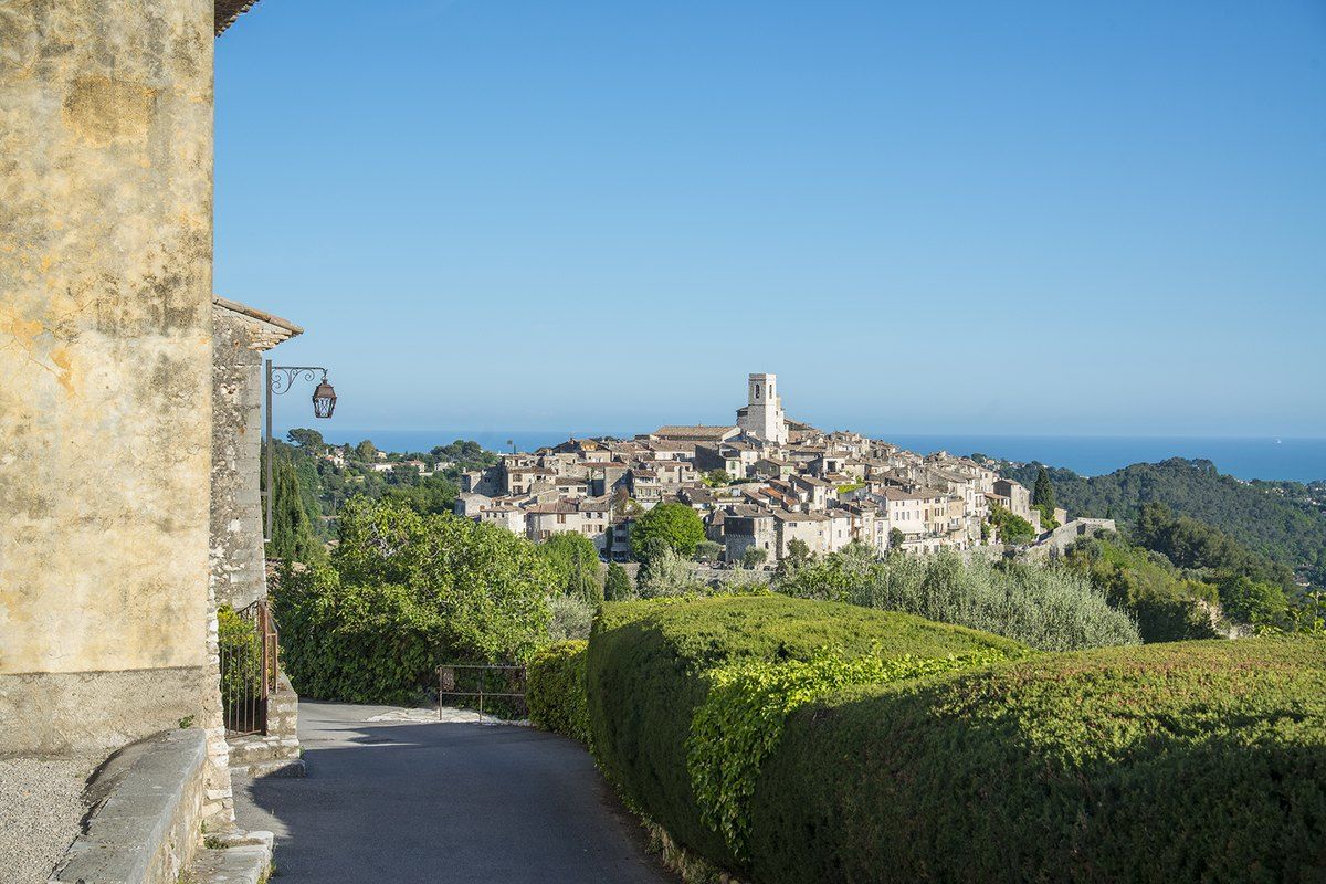 A lane between clipped hedges looking back to the walled village of Saint-Paul de Vence and the sea