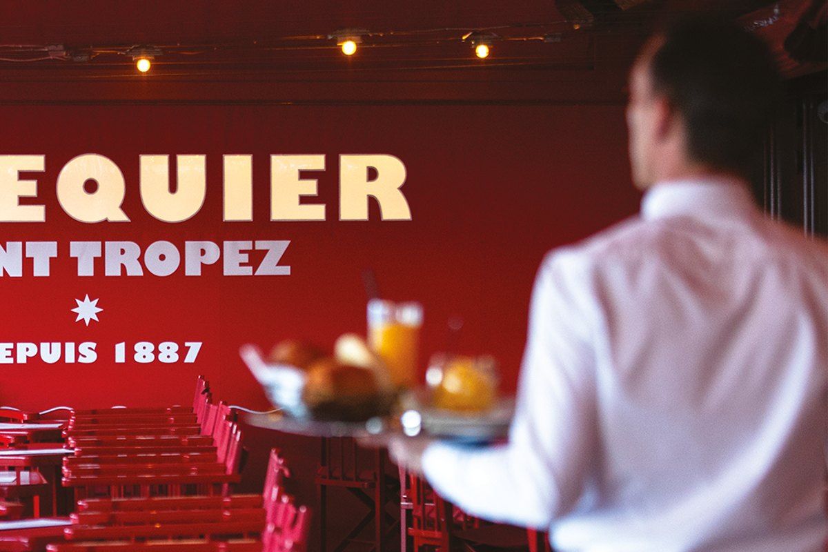 A waiter carrying a tray past the red 'Sénéquier Saint-Tropez depuis 1887' wall and red chairs