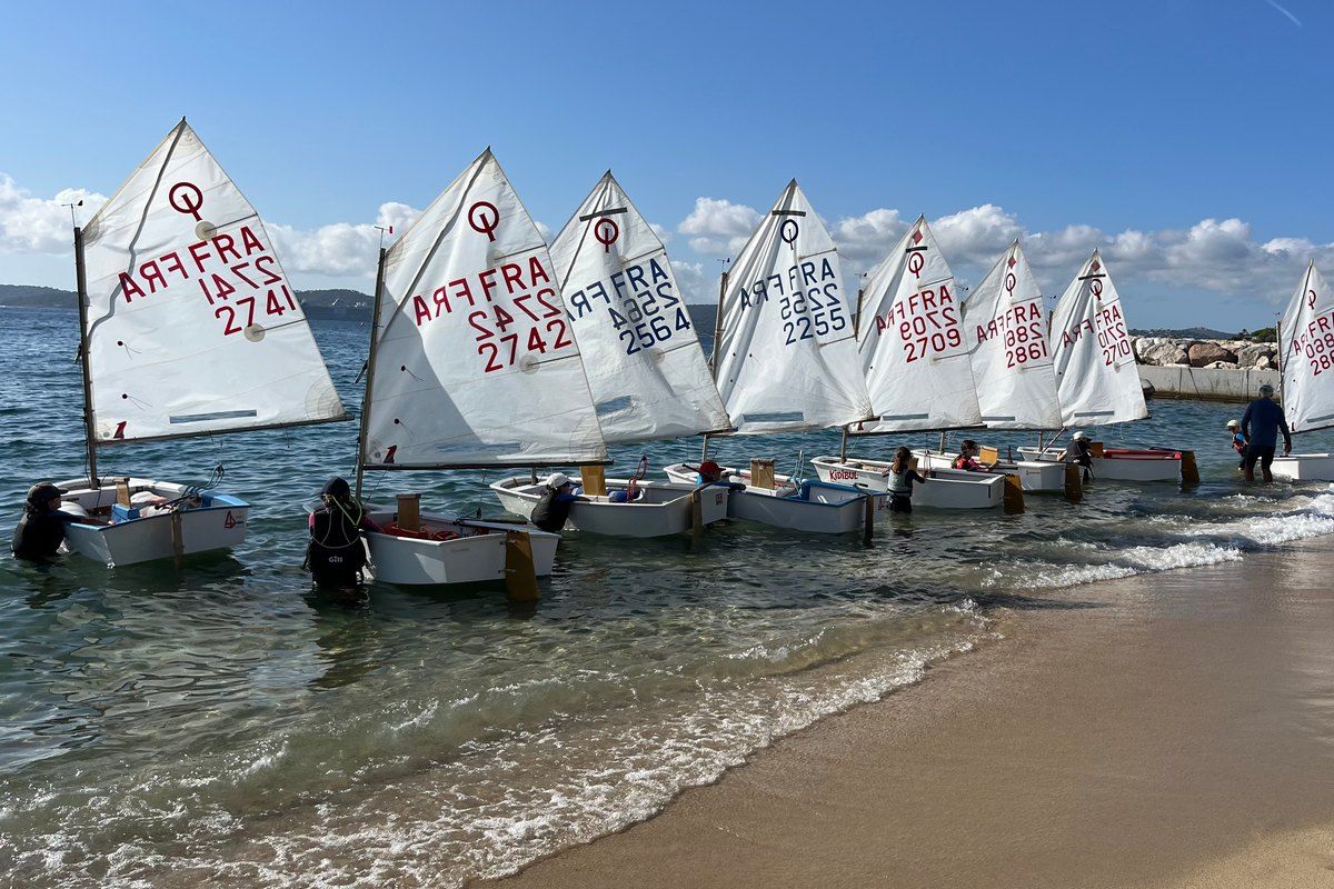 A row of Optimist dinghies at the water's edge in front of the Club Nautique de Sainte-Maxime