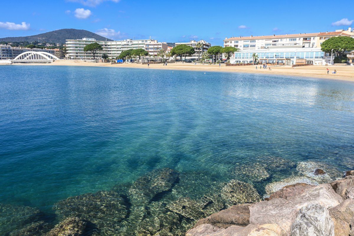 Clear shallow water over rocks at Plage Luc Provensal, with the seafront of Sainte-Maxime and the Préconil footbridge behind