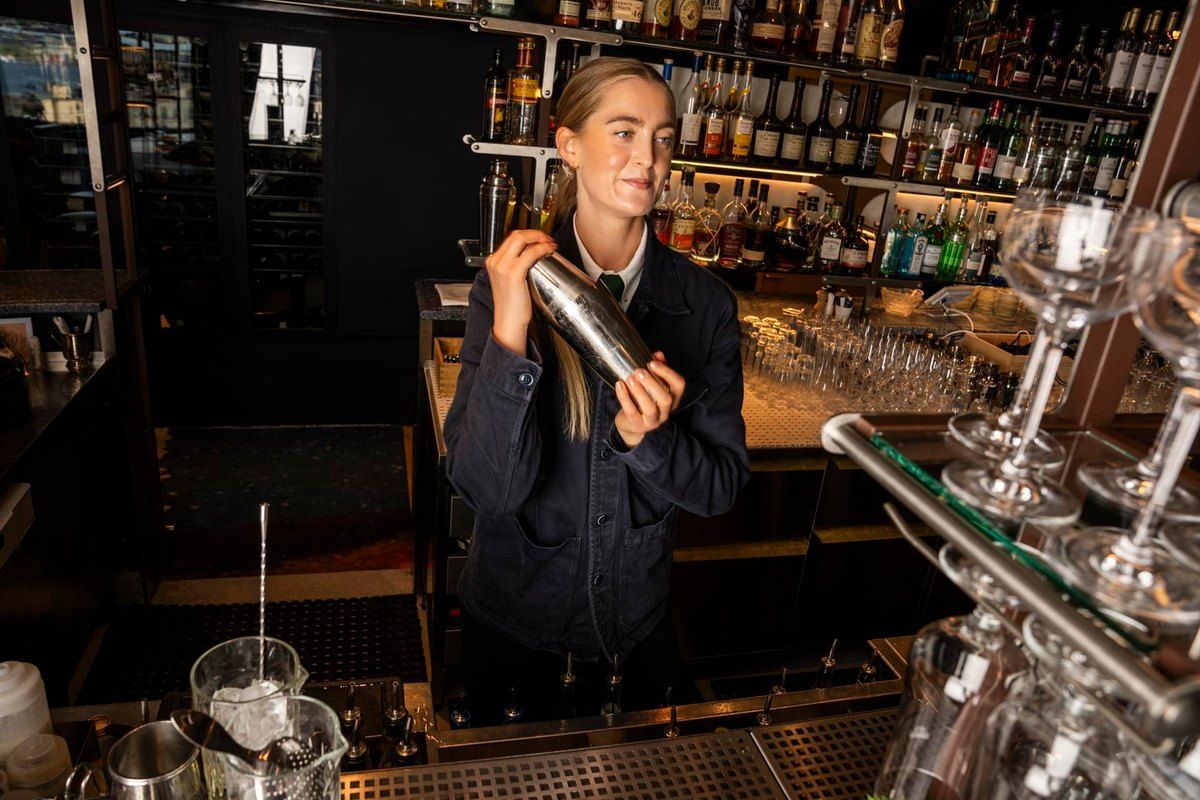 A bartender shaking a cocktail behind the long bar at Gondolen, with lit bottle shelves behind her
