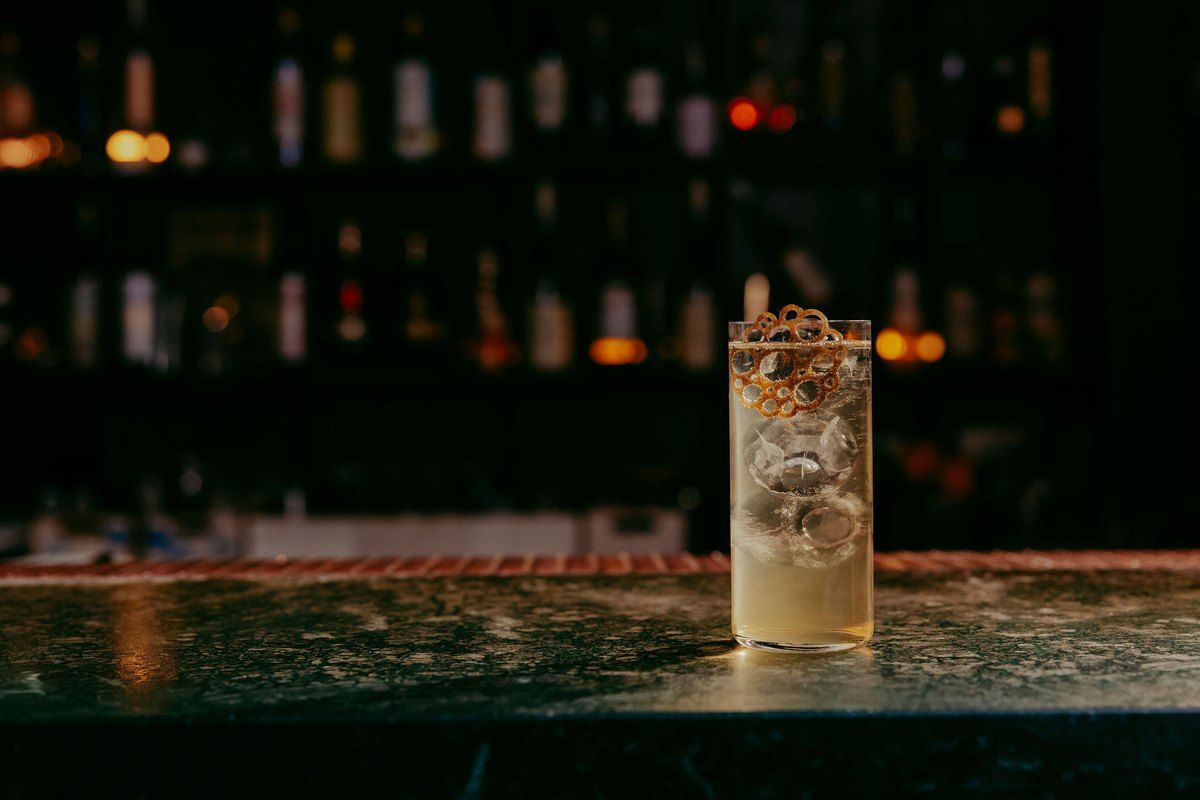 A tall cocktail on ice on the green marble bar at Pharmarium, with bottle shelves glowing behind