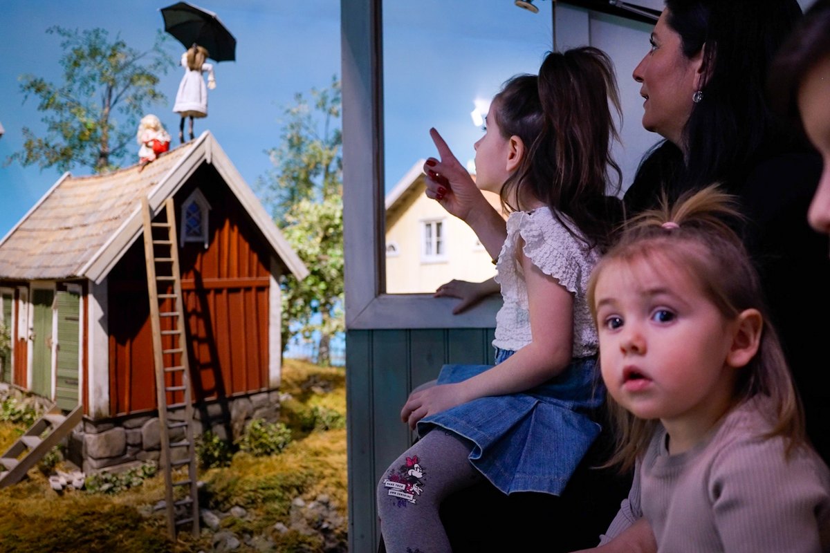 Two young girls watch from the Story Train at Junibacken as it passes a miniature red cottage