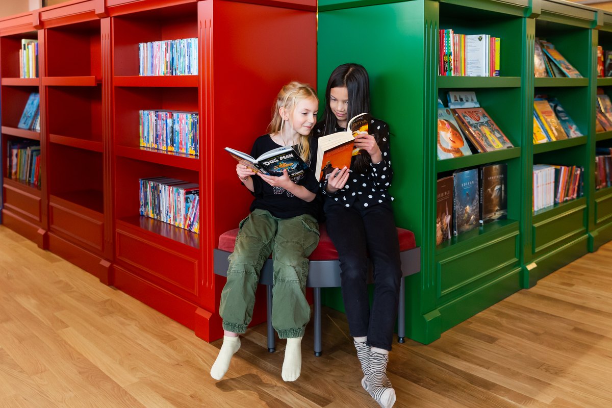 Two girls reading together between red and green bookshelves in the children's library Rum för Barn at Kulturhuset