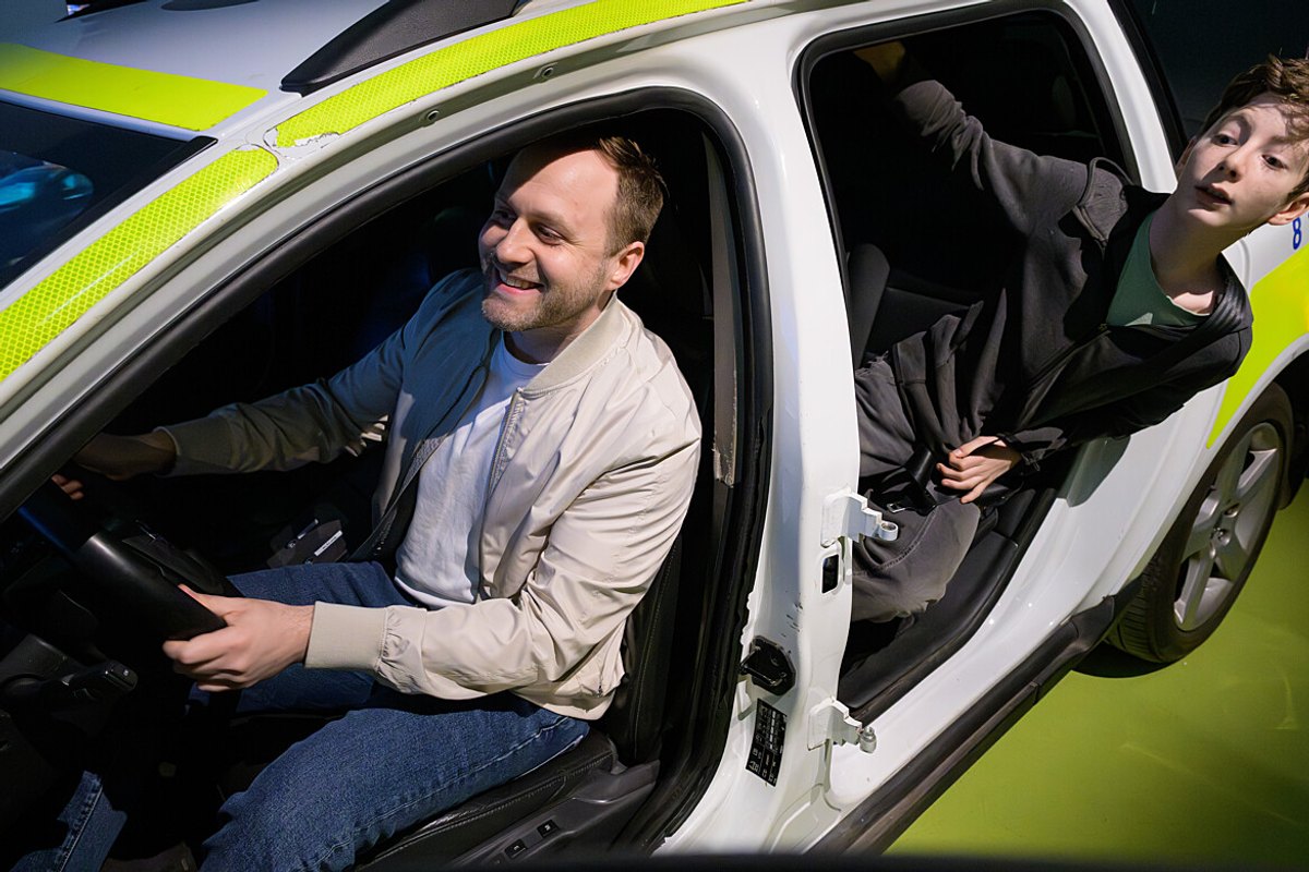 A man at the wheel and a boy leaning out of the back door of a modern police car at Polismuseet