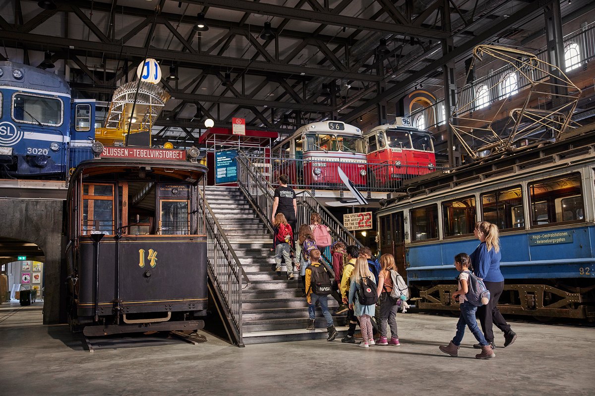 Visitors walking among vintage trams and metro cars in the great hall of Spårvägsmuseet at Gasverket