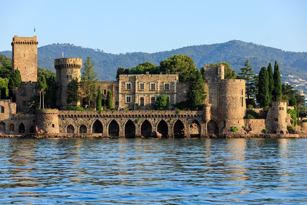 The towers and arcaded sea wall of the Château de La Napoule reflected in calm water, with the Tanneron hills behind