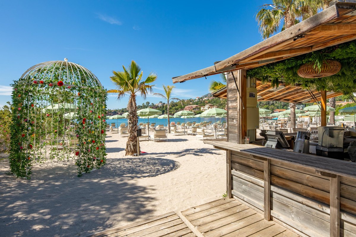 A wooden beach bar and a flower-covered arbour on the sand at La Joya Beach, with palms and the Estérel hills behind