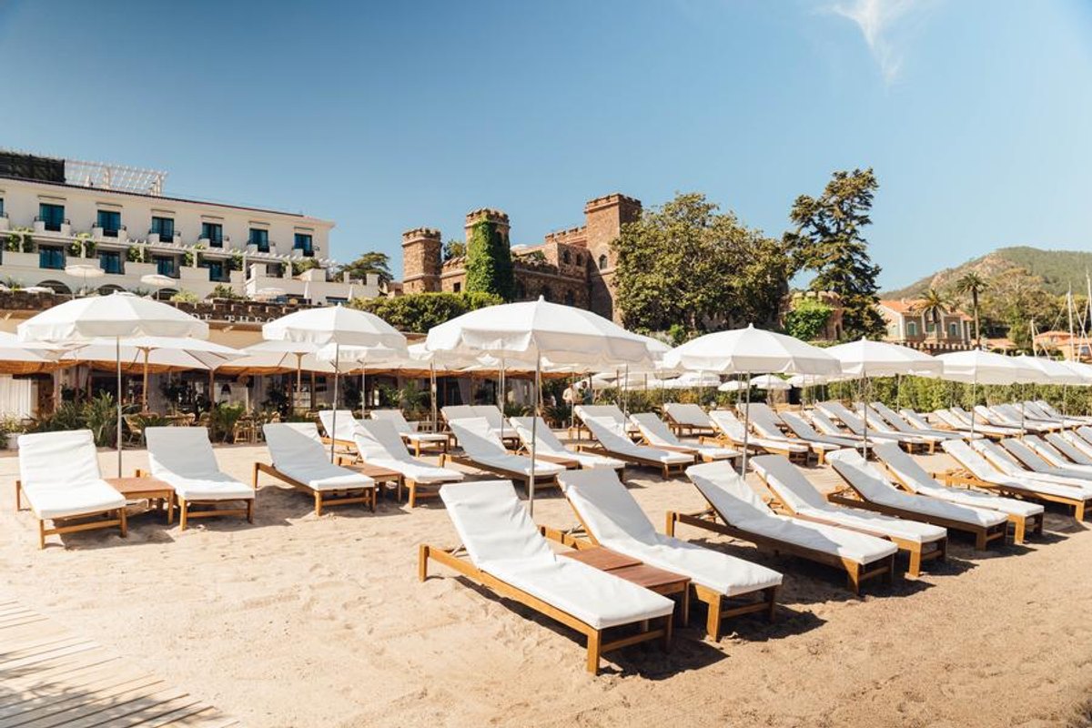 White loungers and parasols on the sand of La Plage Blanche, with the Château de Théoule's towers behind