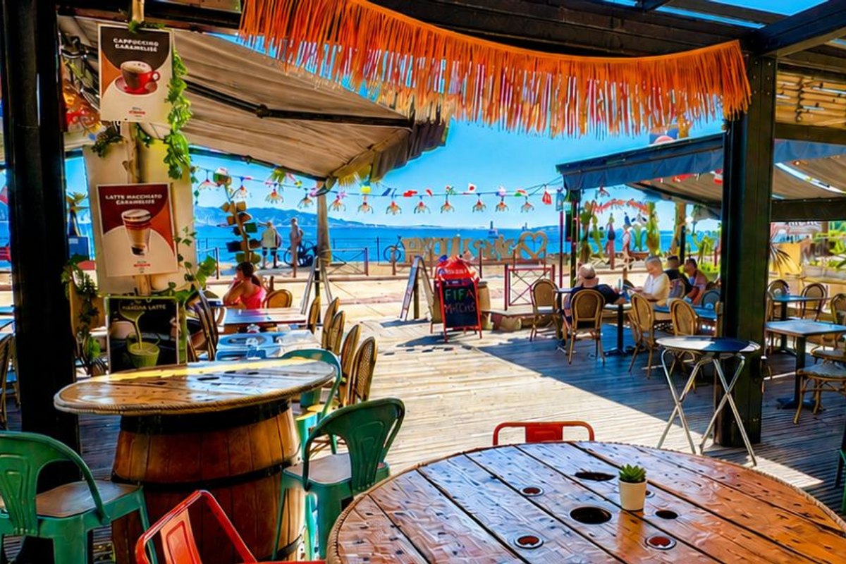 Barrel tables and bright chairs under a fringed awning at La Terrasse de Théoule, looking out to the sea
