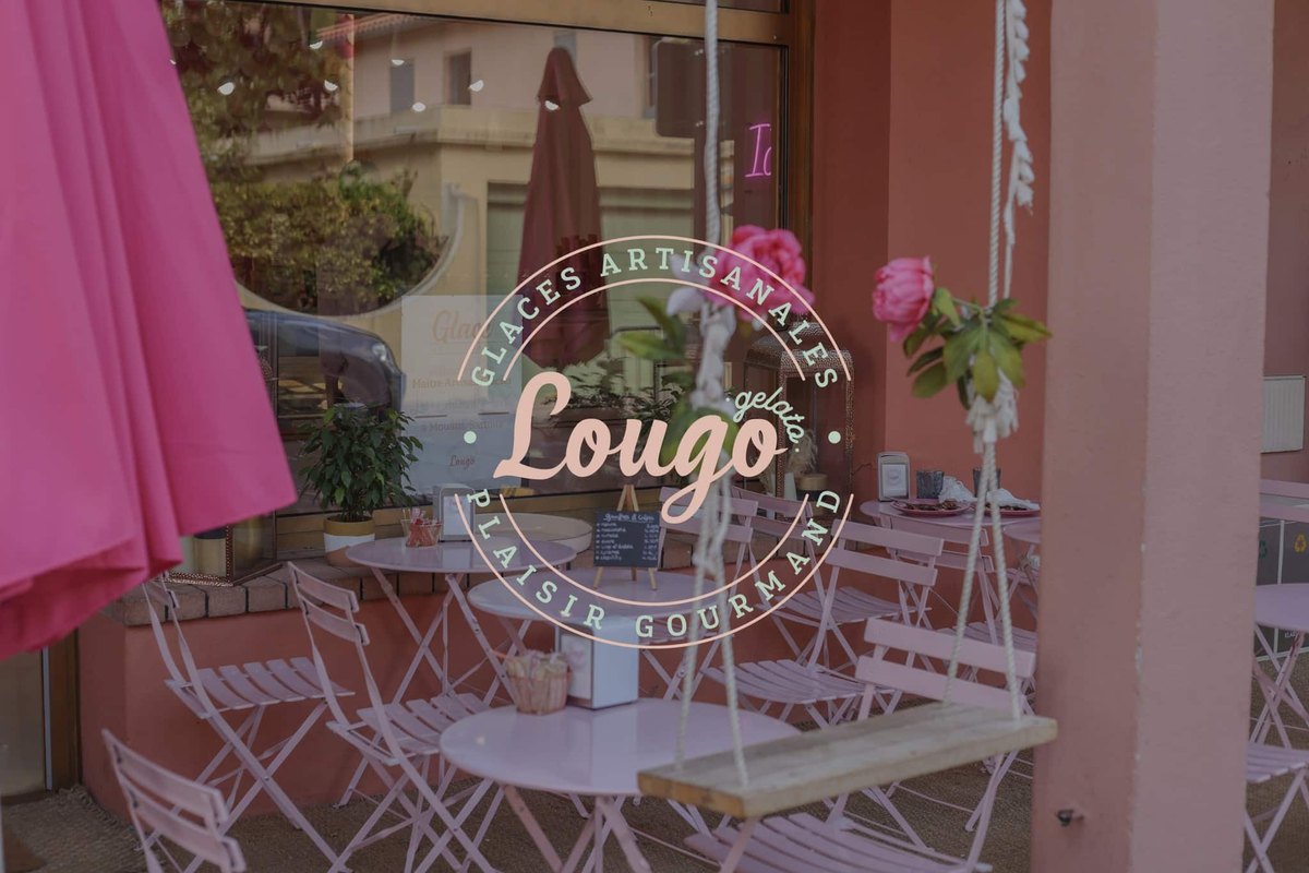 The pink-walled terrace of Lougo Gelato in Théoule, with white café tables, a parasol and the ice-cream shop's logo on the window