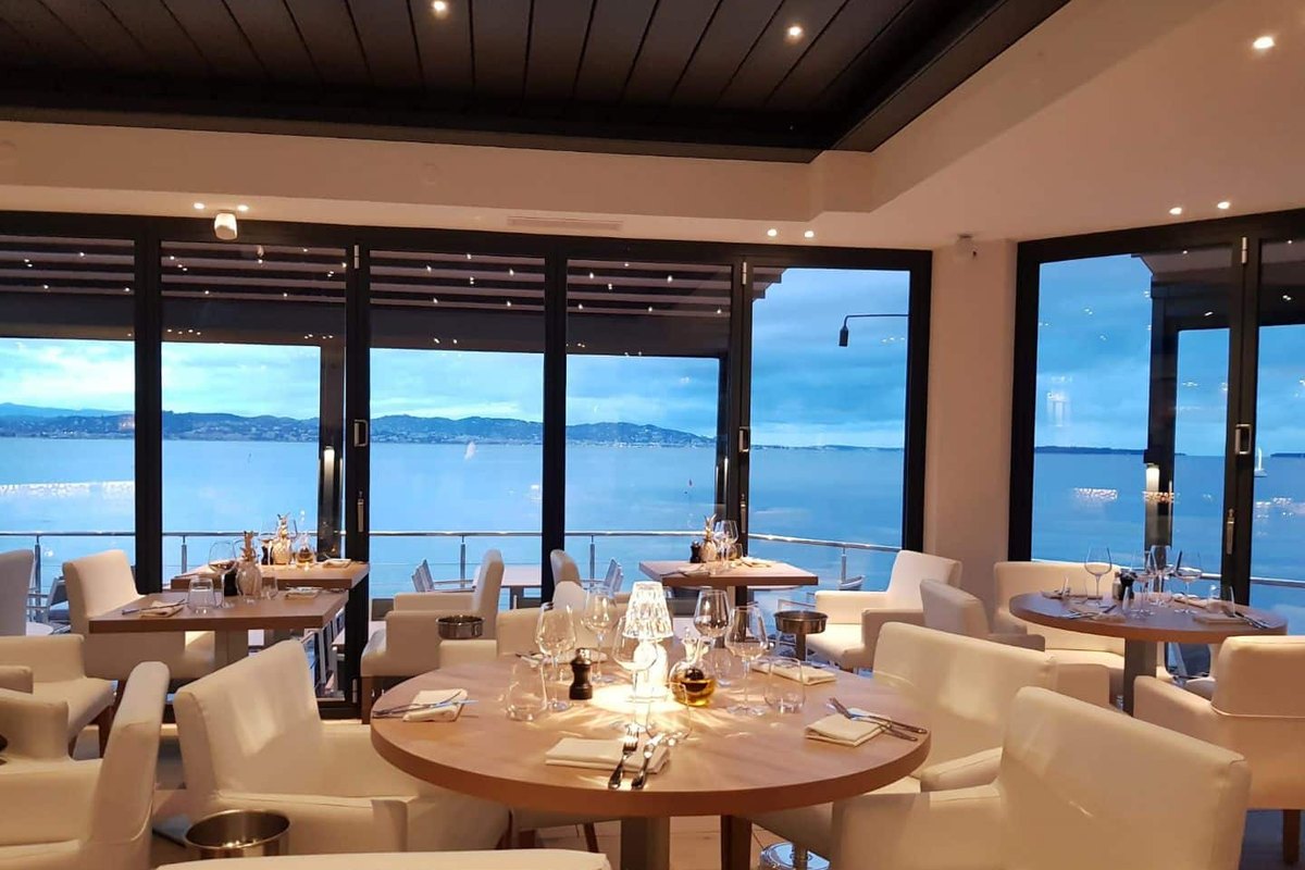 The dining room of Le Marco Polo with round tables and white chairs behind tall windows open onto the bay