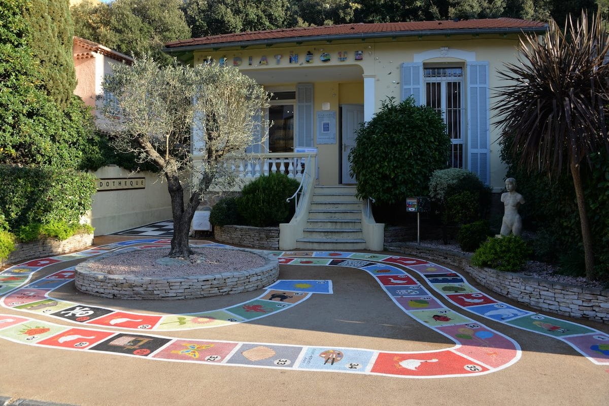 The giant goose game painted on the forecourt of the Villa La Paix médiathèque, with an olive tree and palm in front of the pale yellow villa