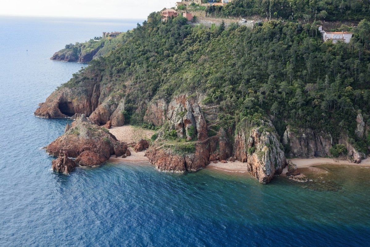 Aerial view of the red-rock coves and small beaches of the Pointe de l'Aiguille below a wooded headland
